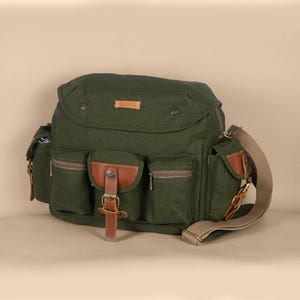Pode incluir: Uma bolsa mensageiro de lona verde-oliva escura com vários bolsos externos com detalhes em couro marrom, uma aba central com fecho de fivela, uma alça de ombro ajustável bege e uma pequena etiqueta retangular com a inscrição DIVINCES.
