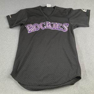 May include: A black V-neck jersey with short sleeves and a mesh texture. The word ROCKIES is printed across the chest in purple and white lettering. A small Majestic logo is visible on the right sleeve and a patch is on the left sleeve.
