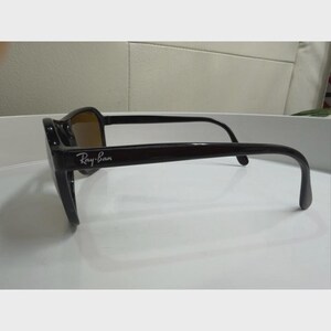 Puede incluir: Gafas de sol de color marr&oacute;n oscuro con acabado brillante y montura rectangular. Las patillas presentan el logotipo de Ray-Ban en blanco impreso en el lateral. Las lentes tienen un tono &aacute;mbar o marr&oacute;n c&aacute;lido, visible de perfil.