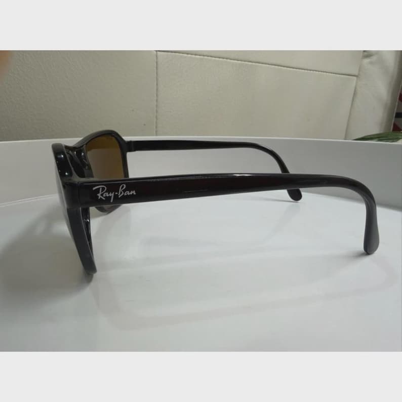 Puede incluir: Gafas de sol de color marr&oacute;n oscuro con acabado brillante y montura rectangular. Las patillas presentan el logotipo de Ray-Ban en blanco impreso en el lateral. Las lentes tienen un tono &aacute;mbar o marr&oacute;n c&aacute;lido, visible de perfil.