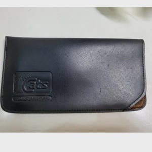 Puede incluir: Un estuche para gafas rectangular de color negro con acabado texturizado. La parte frontal presenta un logotipo en relieve que dice Ray-Ban Cats y Bausch & Lomb. El estuche tiene esquinas redondeadas y una secci&oacute;n de material marr&oacute;n abajo.