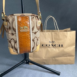 Könnte beinhalten: Eine beige-braune Coach-Umhängetasche mit charakteristischem Monogramm-Canvas-Muster, einem zentralen vertikalen Paneel in orangefarbener Schlangenlederoptik, goldfarbenen Beschlägen und einer passenden braunen Papiertüte mit Coach New York Logo.