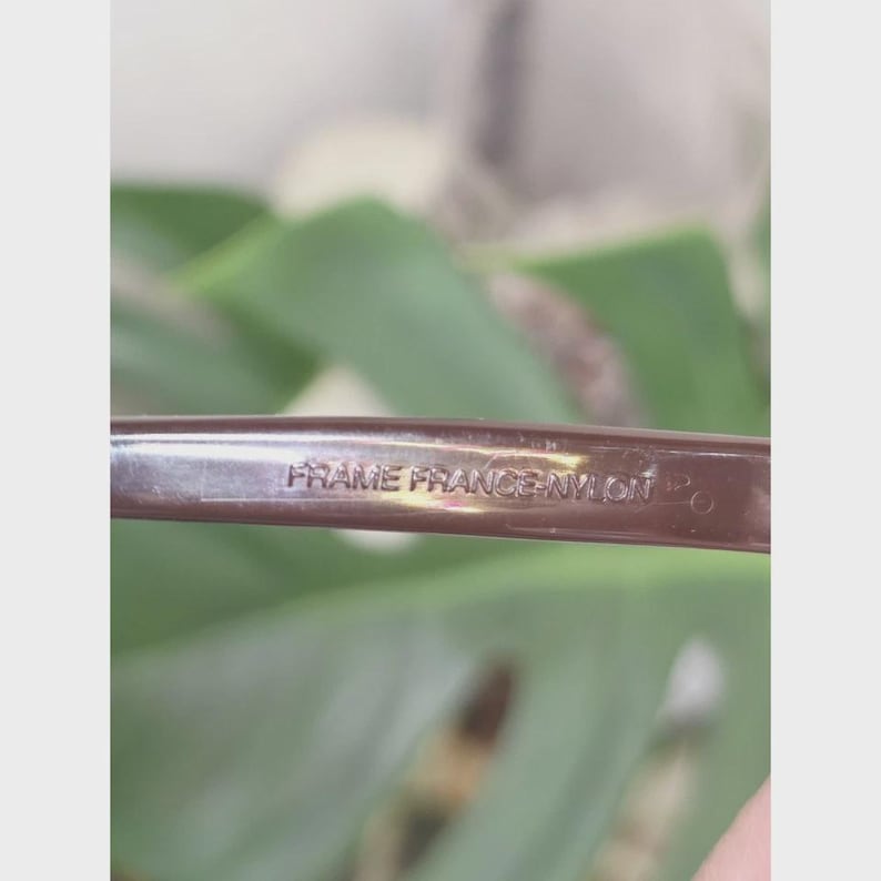 Puede incluir: Una patilla de gafas marr&oacute;n que presenta el texto en relieve FRAME FRANCE NYLON. El material tiene un acabado liso y brillante con una forma ligeramente curva, mostrando claramente el origen y la composici&oacute;n del material.