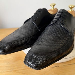 May include: A pair of black lace-up dress shoes featuring a textured snakeskin-patterned vamp and a smooth leather quarter. The shoes have a square toe design and include light-coloured wooden shoe trees with gold-toned metal knobs inserted inside.