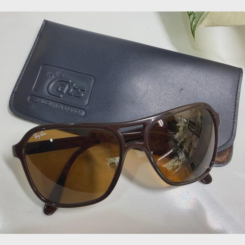 Puede incluir: Gafas de sol de color marr&oacute;n oscuro con doble puente y lentes tintadas en &aacute;mbar, con el logotipo de Ray-Ban en la lente superior derecha. Incluye un estuche protector azul marino con el texto Ray-Ban Cats y Bausch & Lomb en relieve.