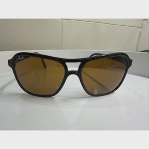 Puede incluir: Gafas de sol con montura negra, dise&ntilde;o de doble puente y lentes tintadas en color marr&oacute;n. La lente izquierda muestra el logotipo de Ray-Ban en letras blancas. La montura tiene un acabado liso y brillante con una barra superior curva.