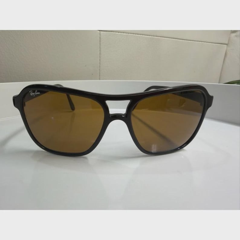 Puede incluir: Gafas de sol con montura negra, dise&ntilde;o de doble puente y lentes tintadas en color marr&oacute;n. La lente izquierda muestra el logotipo de Ray-Ban en letras blancas. La montura tiene un acabado liso y brillante con una barra superior curva.