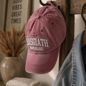 May include: A dusty rose baseball cap featuring the embroidered white text BASGIATH WAR COLLEGE on the front panel. The cap has a structured crown, a curved brim, and a button at the top, hanging from a wooden hook.