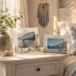 May include: Two rectangular white picture frames decorated with nautical elements including miniature sailboats, life preservers, fishing nets, and various seashells. The frames hold photographs of a coastal scene and a sailboat on the water.