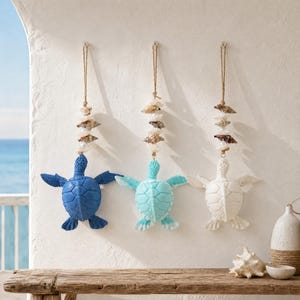 May include: Three hanging sea turtle ornaments in blue, light blue, and white. Each turtle is suspended by a jute rope adorned with various small seashells and white beads, creating a vertical display of marine-themed wall decor.