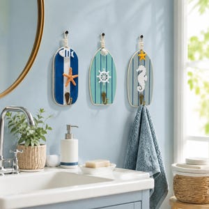 May include: Three surfboard-shaped wall hooks with nautical motifs. The first is blue and white with a fish and starfish. The second is teal with a ship wheel. The third is grey with a yellow starfish and white seahorse, holding a blue towel.