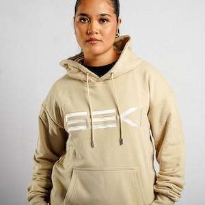 May include: A beige hooded sweatshirt featuring a front kangaroo pocket and long sleeves. The chest displays the word SEKI in white, stylised, horizontal-lined lettering. The hood includes matching beige drawstrings with metallic silver tips.