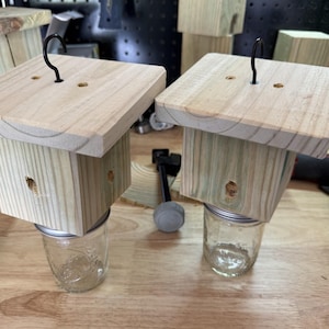 May include: Two square wooden blocks with flat tops and small black metal hooks attached to the center. Each block is mounted onto a clear glass jar lid, with the glass jar body extending downward. The wood has a light natural grain finish.