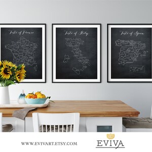 May include: Three black framed prints of maps of France, Italy, and Spain with the text "Taste of France", "Taste of Italy", and "Taste of Spain" above each map. The maps are on a dark gray background with white lines outlining the regions. The prints are hanging on a white wall above a wooden table with a vase of sunflowers, a bowl of fruit, and two white chairs.