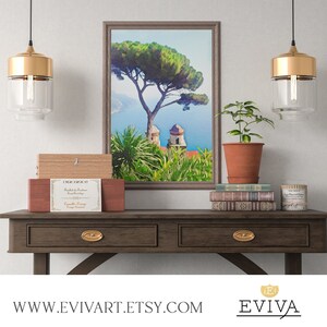 May include: A wooden table with two drawers and gold-toned hardware. The table is decorated with a framed painting of a tree and a building, a potted plant, a stack of books, and a candle. The painting is a watercolour style with a blue sky and green trees. The table is in front of a white wall.