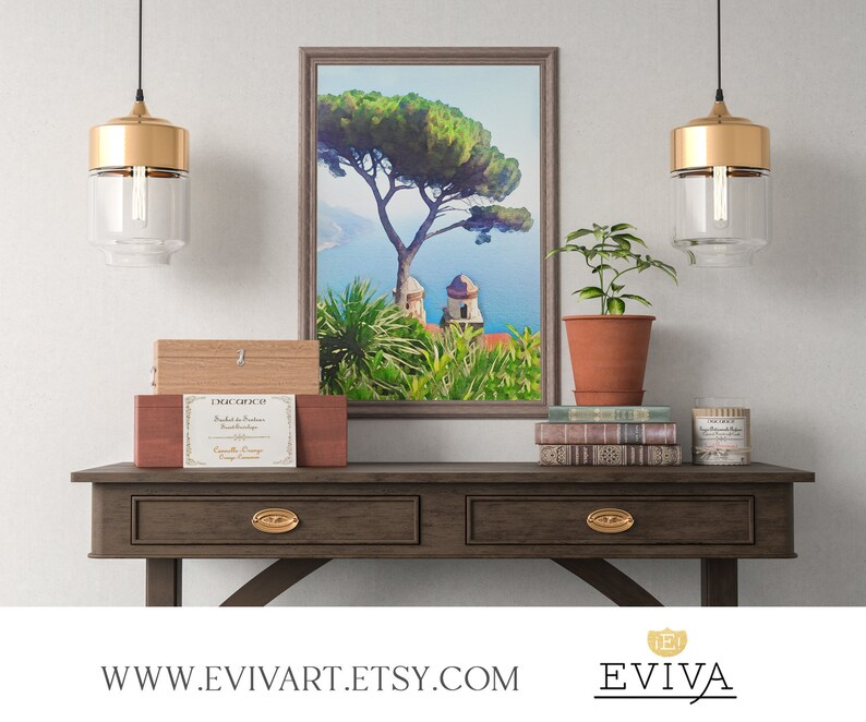 May include: A wooden table with two drawers and gold-toned hardware. The table is decorated with a framed painting of a tree and a building, a potted plant, a stack of books, and a candle. The painting is a watercolour style with a blue sky and green trees. The table is in front of a white wall.