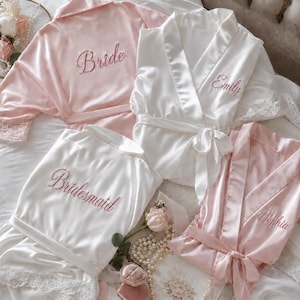 Puede incluir: Cuatro batas de satén en rosa claro y blanco, con ribetes de encaje en las mangas y los dobladillos. Las batas están bordadas con las palabras Bride, Emily, Bridesmaid y Sophia en letra cursiva rosa. Se incluyen un joyero rosa y perlas.