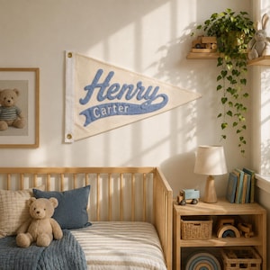 以下が含まれることがあります： 青い筆記体でHenry Carterという名前が刺繍された、クリーム色の三角形のフェルト製ペナント旗。左側には吊り下げ用の金色の金属製ハトメが2つ付いており、木製のベビーベッドの上の壁に飾られています。
