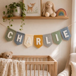 May include: A felt name banner spelling CHARLIE with individual pennant flags in muted shades of sage green, slate blue, cream, mustard yellow, and gray. Each flag features a large, white, capital letter stitched onto the center of the fabric.
