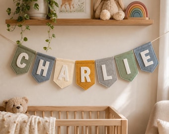 Può includere: Uno striscione in feltro con il nome CHARLIE, composto da bandierine individuali in tonalità tenui di verde salvia, blu ardesia, crema, giallo senape e grigio. Ogni bandierina presenta una grande lettera maiuscola bianca cucita al centro.