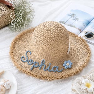 May include: A tan straw sun hat with a frayed edge, featuring the name Sophia embroidered in blue yarn script across the front brim, accented by a small, light blue crocheted flower attached to the right side of the brim.