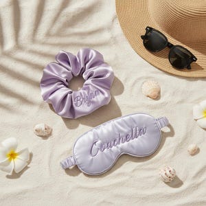 May include: A lavender satin scrunchie embroidered with the name Bryna and a matching lavender satin sleep mask embroidered with the word Coachella, accompanied by a straw hat, black sunglasses, white flowers, and small seashells.