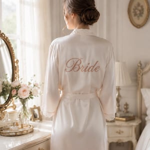May include: A champagne-coloured satin robe featuring long sleeves with delicate lace trim at the cuffs. The back of the robe is embroidered with the word Bride in a cursive script. The garment includes a matching satin tie belt at the waist.