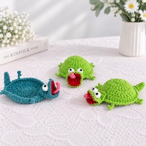 May include: Three crocheted turtle-shaped coasters. One is teal, and two are bright green. Each turtle features a wide-open red mouth, a pink tongue, and two large white eyes with black pupils. The coasters have textured shells and small legs.