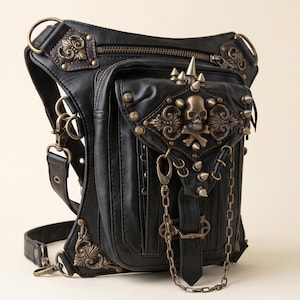 May include: A black leather drop leg bag featuring antique bronze hardware, including a skull and crossbones emblem, metal studs, and a decorative chain. The bag includes multiple zippered compartments, adjustable straps, and ornate metal corner accents.