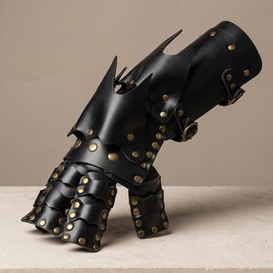 May include: A black leather gauntlet featuring articulated finger plates and a protective wrist section. The piece is constructed with multiple overlapping segments secured by metallic brass-colored rivets and includes adjustable buckle straps.