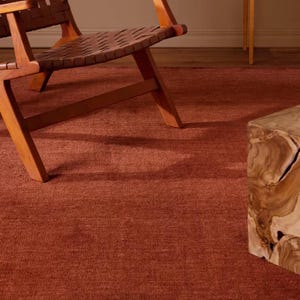 Könnte beinhalten: Ein einfarbiger, terrakottafarbener Teppich mit einer strukturierten, gewebten Oberfläche. Der Teppich liegt unter einem Holzstuhl mit geflochtener Sitzfläche und einem Beistelltisch aus Naturholz mit markanter Maserung und organischen Kanten.
