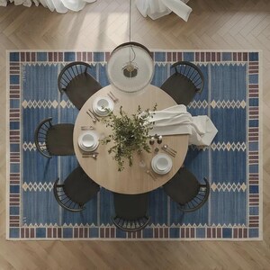 May include: A round light wood dining table surrounded by six dark wood spindle chairs, set upon a large blue area rug with a geometric border pattern. The table is arranged with white plates, silverware, and a central green plant centerpiece.
