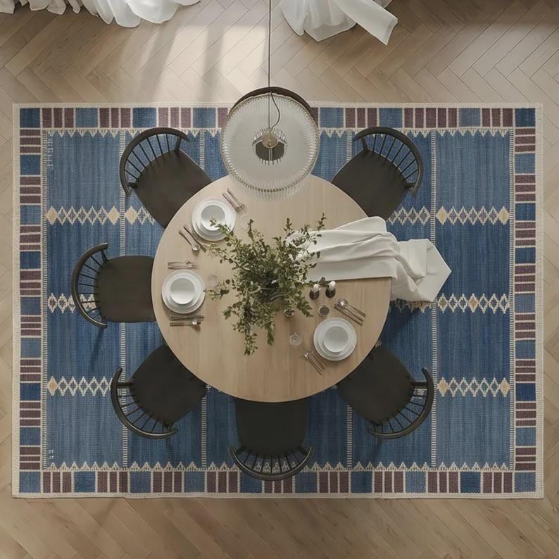 May include: A round light wood dining table surrounded by six dark wood spindle chairs, set upon a large blue area rug with a geometric border pattern. The table is arranged with white plates, silverware, and a central green plant centerpiece.