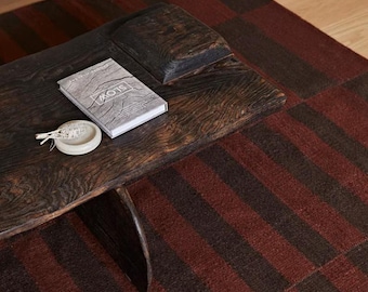 May include: A dark brown wooden coffee table with a live edge, featuring a rectangular top and a curved support base. A book with the word SLOW on the cover and a small, round, off-white ceramic dish containing dried herbs sit on the table surface.