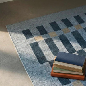 May include: A rectangular area rug featuring a geometric pattern of dark blue, light blue, and beige rectangles. A stack of four books with blue, tan, green, and orange covers rests on the rug, which has a textured, woven appearance.