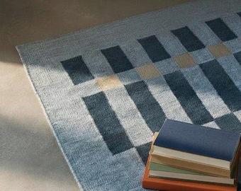 May include: A rectangular area rug featuring a geometric pattern of dark blue, light blue, and beige rectangles. A stack of four books with blue, tan, green, and orange covers rests on the rug, which has a textured, woven appearance.