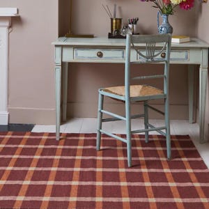 May include: A rectangular area rug featuring a woven plaid pattern in shades of deep red, burnt orange, and cream. The rug is positioned on a white floor beneath a light blue wooden desk and a matching chair with a woven straw seat.