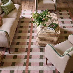 May include: A rectangular area rug featuring a grid pattern of intersecting pink, cream, and dark green stripes. The rug is positioned beneath a light-coloured sofa, two matching armchairs, and a rounded rectangular coffee table with a striped design.