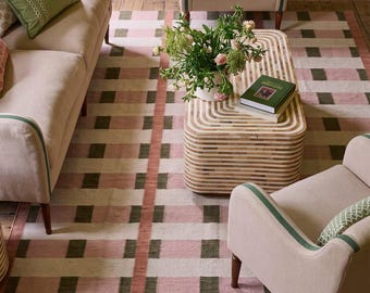 May include: A rectangular area rug featuring a grid pattern of intersecting pink, cream, and dark green stripes. The rug is positioned beneath a light-colored sofa, two matching armchairs, and a rounded rectangular coffee table with a striped design.