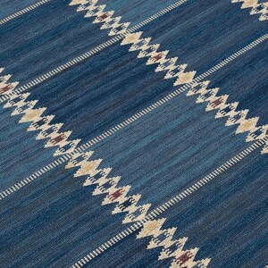 May include: A woven rug featuring a deep blue background with diagonal stripes. The stripes consist of repeating diamond patterns in cream and tan, accented with small dark red details, creating a textured surface with a visible fabric weave.