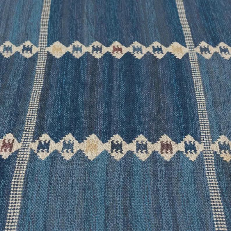 May include: A woven rug featuring a deep blue and teal gradient background with vertical white checkered stripes. Horizontal bands of repeating diamond-shaped motifs in white, tan, and dark brown are spaced evenly across the textured surface.
