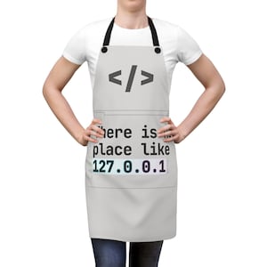 Può includere: Un grembiule grigio chiaro con lacci neri al collo e in vita. Il grembiule presenta un simbolo di parentesi di codice grigio scuro in alto e il testo "There is no place like 127.0.0.1" stampato in nero, con l'indirizzo IP evidenziato.