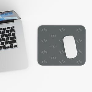 May include: A rectangular dark gray mouse pad featuring a repeating light gray pattern of coding bracket symbols. A white computer mouse rests on the surface of the mouse pad, positioned next to a silver laptop with a black keyboard.