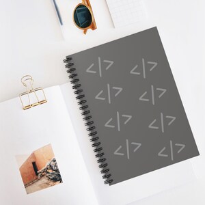 Puede incluir: Un cuaderno de espiral con una cubierta gris oscuro que presenta un patrón repetitivo de símbolos de corchetes de código en gris claro. El cuaderno está abierto, mostrando una página blanca con una pequeña foto cuadrada de un desierto y un clip.