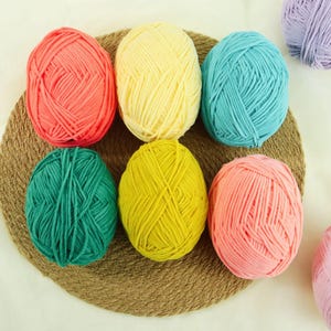 May include: Six oval-shaped balls of yarn in vibrant colours including coral, pale yellow, turquoise, dark green, bright yellow, and light pink, arranged in two rows on a round, textured, woven brown mat, with a partial lavender ball visible.