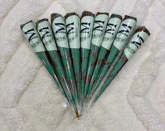 May include: Nine cone-shaped henna applicators arranged in a fan pattern. Each cone is wrapped in light green paper featuring a dark green floral wreath design and the letter M in a serif font. The text Henna by Munifa is printed on the cones.