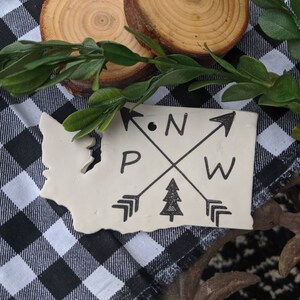Puede incluir: Una pieza de cerámica de color crema con la forma del estado de Washington, con las letras PNW y flechas. El diseño es en tinta negra. La pieza está sobre una superficie a cuadros blancos y negros, con vegetación y rodajas de madera.