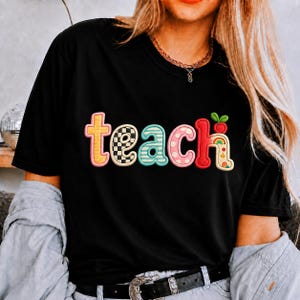 以下が含まれることがあります： 黒の半袖Tシャツで、大きくカラフルな柄入りの文字でteachとプリントされています。各文字は、黄色、白黒の市松模様、ティールブルーのストライプ、ピンクの水玉模様、小さなリンゴが描かれた赤など、ユニークなデザインになっています。