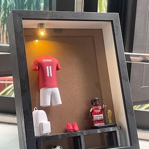 May include: A black shadow box frame containing a red football shirt with the name QUANG and number 11, a white backpack, a pair of red boots, a football, a small figurine in a red shirt, and a gold trophy, illuminated by a small light.