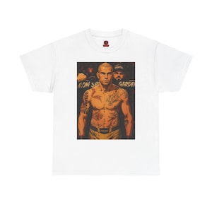 Könnte beinhalten: Ein weißes Kurzarm-T-Shirt mit einem zentralen Grafikdruck eines oberkörperfreien, tätowierten Kämpfers mit kahlgeschorenem Kopf. Der Kämpfer trägt eine gelbe Hose mit dem Wort FIGHTER auf dem Bund. Eine zweite Person ist im Hintergrund zu sehen.